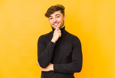 Sarı bir zemin üzerinde izole edilmiş genç bir Arap adam gülümsüyor mutlu ve kendinden emin, elleriyle çenesine dokunuyor..