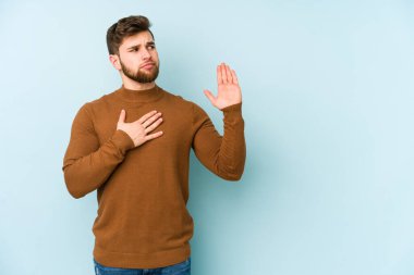 Kafkasyalı genç adam mavi arka planda izole edilmiş yemin ediyor, elini göğsüne koyuyor..