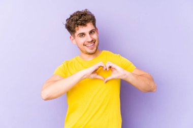 Sarışın, kıvırcık saçlı, beyaz tenli, soyutlanmış, gülümseyen ve elleriyle kalp şeklini gösteren bir adam..