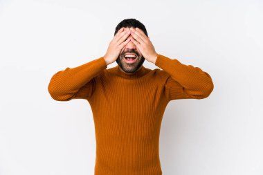 Beyaz bir arka planı olan genç bir latin adam, elleri ile gözlerini kapatıyor, geniş bir gülümsemeyle bir sürpriz bekliyor..