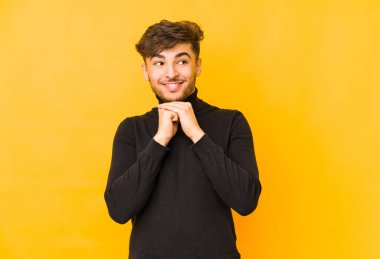 Sarı arka planda izole edilmiş genç Arap adam ellerini çenesinin altında tutuyor ve mutlu bir şekilde yan tarafa bakıyor..