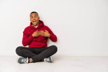 Yerde tek başına oturan genç latin adam gülüyor, elini yüreğine koyuyor, mutluluk kavramını koruyor..