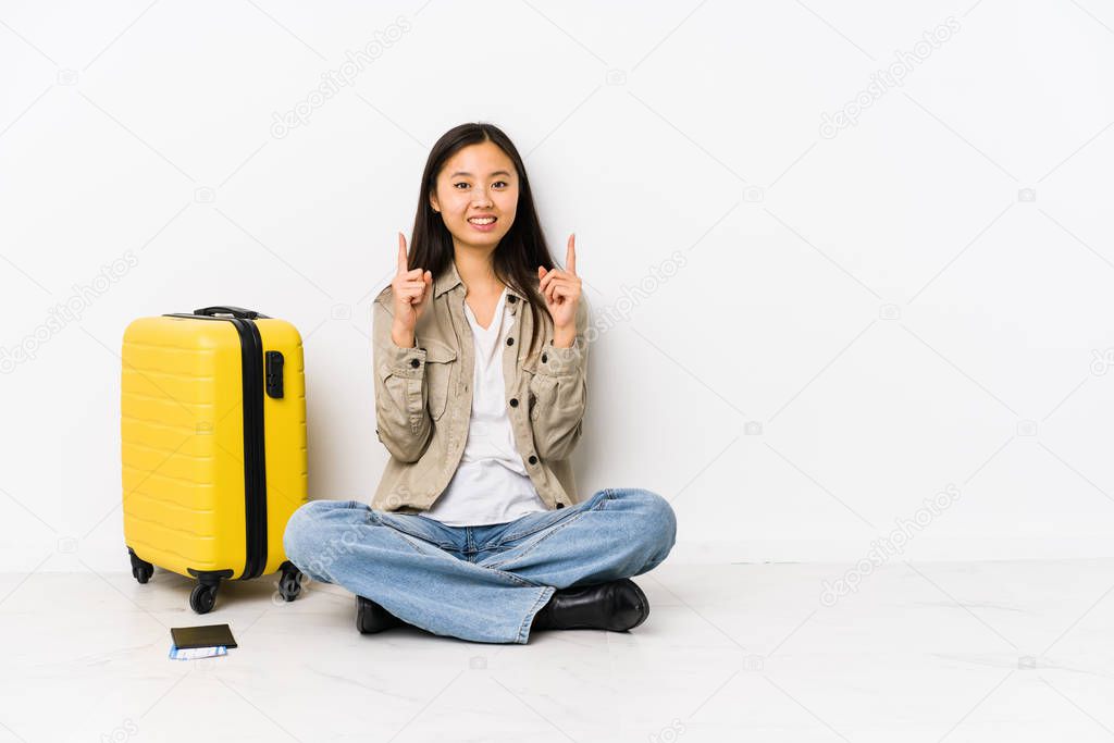 Young chinese traveler woman sitting holding a boarding passes indicates with both fore fingers up showing a blank space.