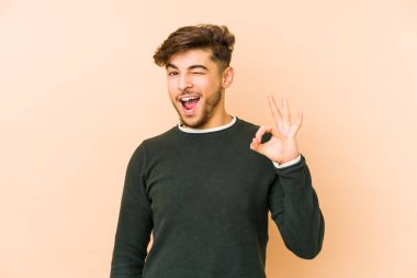 Genç Arap bir adam bej bir arka planda izole edilmiş bir şekilde göz açıp kapayıncaya kadar iyi bir el hareketi yapıyor..