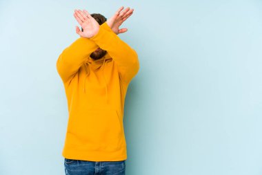 Kafkasyalı genç adam mavi arka planda izole edilmiş iki kolu çapraz tutuyor, inkar kavramı.