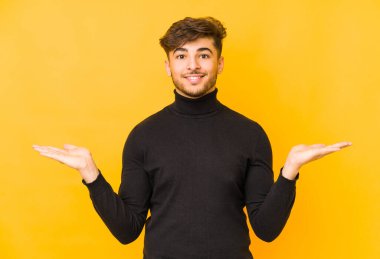 Sarı arka planda izole edilmiş genç bir Arap adam kollarıyla ölçek oluşturuyor, mutlu ve kendinden emin hissediyor..