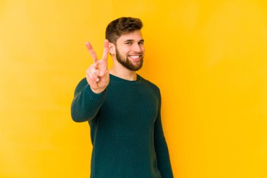 Sarı arka planda izole edilmiş genç beyaz adam zafer işareti gösteriyor ve gülümsüyor..