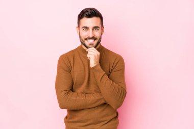 Pembe arka planda genç beyaz bir adam izole edilmiş mutlu ve kendinden emin gülümsüyor, elleriyle çenesine dokunuyor..