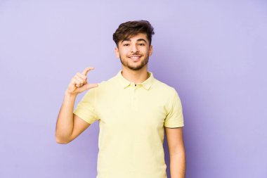 Mor arka planda izole edilmiş genç bir Arap adam işaret parmaklarıyla küçük bir şey tutuyor, gülümsüyor ve kendinden emin..