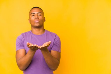 Sarı arka planda izole edilmiş genç latin adam dudakları katlıyor ve hava öpücüğü göndermek için avuç içleri tutuyor..