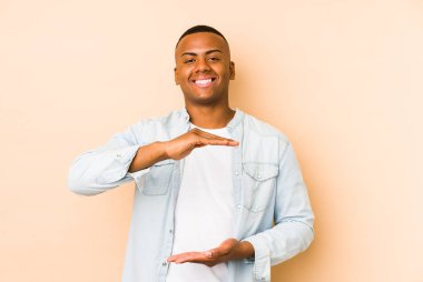 Genç latin adam bej arka planda izole edilmiş iki eliyle bir şey tutuyor, ürün sunumu..