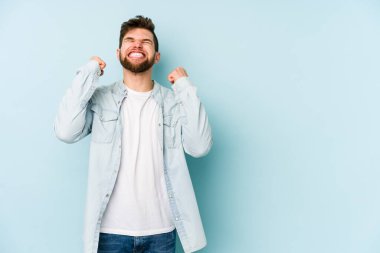 Mavi arka planda izole edilmiş genç beyaz bir adam zaferi, tutkuyu ve coşkuyu kutluyor, mutlu bir ifade..