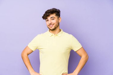 Mor arka planda izole edilmiş genç bir Arap adam ellerini kalçasında tutuyor..