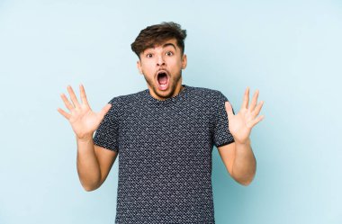 Genç Arap bir adam mavi bir arkaplanda zaferi ya da başarıyı kutlarken şaşırır ve şok olur..