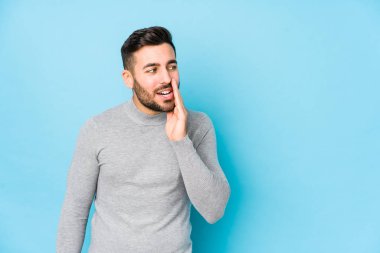 Young caucasian man against a blue background isolated is saying a secret hot braking news and looking aside