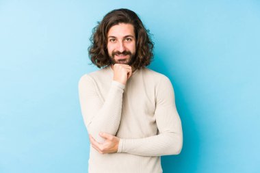 Uzun saçlı genç adam mavi bir arka planda izole edilmiş mutlu ve kendinden emin gülümsüyor, elleriyle çenesine dokunuyor..