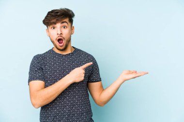 Mavi arka planda izole edilmiş genç bir Arap, palmiyesinde bir kopya alanı tutarken heyecanlanıyor..