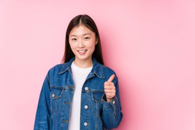 Pembe arka planda poz veren Çinli genç bir kadın yalnız, gülümseyen ve baş parmağını kaldıran.