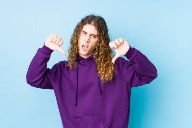 Uzun saçlı adam yalnız poz veriyor, baş parmağını aşağı gösteriyor ve hoşlanmadığını söylüyor..