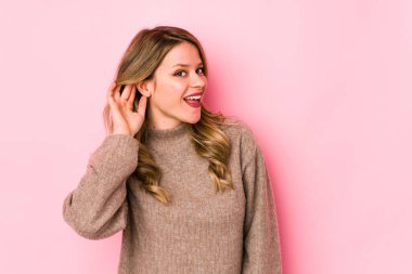 Pembe arka planda izole edilmiş genç beyaz kadın bir dedikoduyu dinlemeye çalışıyor..