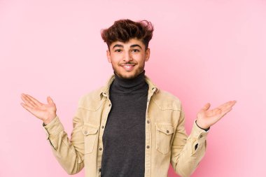 Arka planda yalnız poz veren genç bir Arap adam kollarıyla ölçek oluşturuyor, mutlu ve kendinden emin hissediyor..