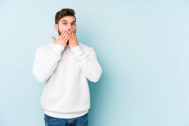 Mavi arka planda izole edilmiş genç beyaz adam elleriyle ağzını kapatırken şok oldu..