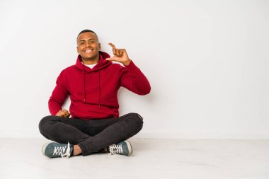 Yerde tek başına oturan genç latin adam işaret parmağıyla küçük bir şey tutuyor, gülümsüyor ve kendinden emin..