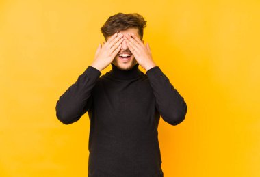 Sarı arka planda izole edilmiş genç bir Arap adam elleriyle gözlerini kapatıyor, geniş bir gülümsemeyle bir sürpriz bekliyor..