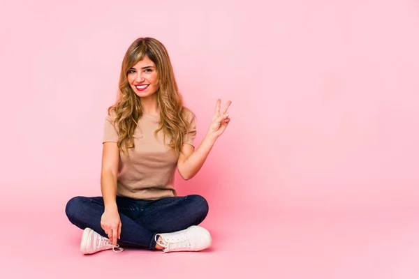 若いですブロンドCaucasian女性座っています上のピンクのスタジオショー勝利サインと笑顔広く — ストック写真