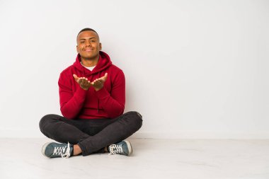 Yerde tek başına oturan genç latin adam elinde avuç içi bir şeyle kameraya sunuyor..