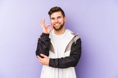 Mor arka planda izole olmuş genç bir adam bir göz kırpıyor ve elleriyle iyi bir jest yapıyor..