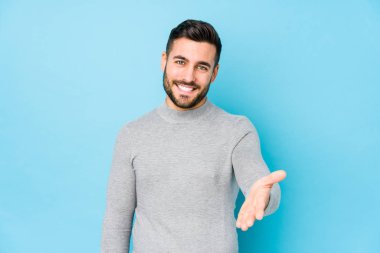 Mavi arka planda genç beyaz bir adam yalnız başına kameraya doğru elini uzatarak selam veriyor..