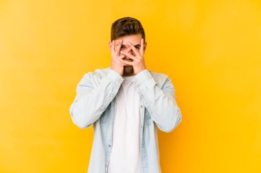Sarı arka planda izole edilmiş genç beyaz adam kameraya parmaklarıyla göz kırpıyor, yüzünü kapatmaktan utanıyor..