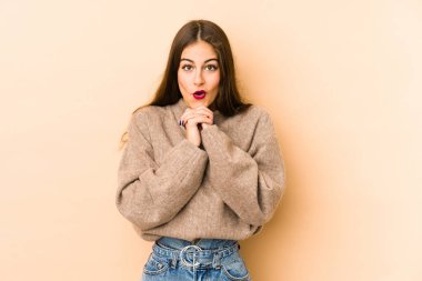 Beyaz tenli genç bir kadın bej arka planda izole edilmiş şans için dua ediyor, şaşırmış ve ağzını açıp önüne bakıyor..