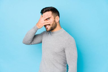 Mavi arka planda genç beyaz bir adam kameraya parmaklarıyla göz kırpıyor, yüzünü kapatmaktan utanıyor..