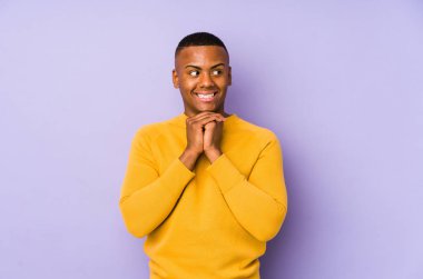 Mor arka planda izole edilmiş genç latin adam ellerini çenesinin altında tutuyor, mutlu bir şekilde yana bakıyor..