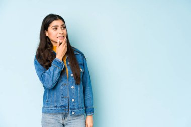 Genç Hintli kadın mavi arka planda izole edilmiş kafasının arkasına dokunuyor, düşünüyor ve bir seçim yapıyor..