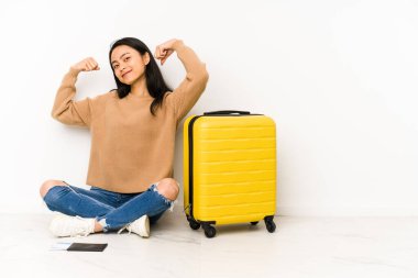 Genç Çinli gezgin kadın, elinde bir bavulla yerde oturuyor. Kollarıyla güçlü bir hareket sergiliyor. Kadınsı gücün sembolü.