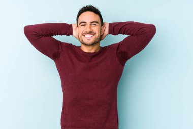 Kafkasyalı genç adam mavi arka planda izole edilmiş. Kendine güveniyor, elleri başının arkasında..