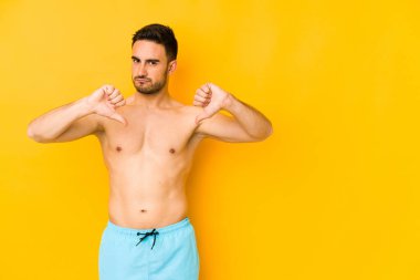 Sarı arka planda mayosu olan genç, beyaz bir adam. Başparmağını aşağı çeken beyaz bir adam. Hayal kırıklığı konsepti..