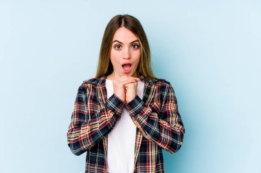 Kafkasyalı genç bir kadın mavi arka planda izole edilmiş şans için dua ediyor, şaşırmış ve ağzını açıp önüne bakıyor..