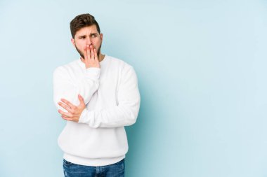 Mavi arka planda izole edilmiş genç beyaz adam esniyor. Ağzı eliyle kapatan yorgun bir hareket sergiliyor..