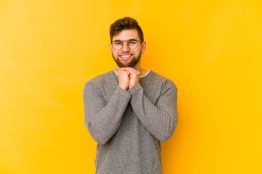 Sarı arka planda izole edilmiş genç beyaz adam ellerini çenesinin altında tutuyor ve mutlu bir şekilde yan tarafa bakıyor..