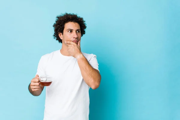 Young Caucasian Curly Man Holding Tea Cup Looking Sideways Doubtful — 스톡 사진