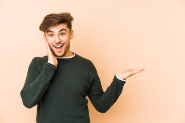 Bej bir arka planda izole edilmiş genç bir Arap, avuç içinde fotokopi alanı tutuyor, el üstünde tutuyor. Şaşırmış ve mutlu..