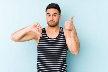 Kafkasyalı genç adam mavi arka planda izole edilmiş, baş parmağını kaldırıyor, zor seçim konsepti.