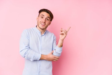 Pembe arka planda poz veren beyaz bir genç yalnız ve neşeli bir şekilde işaret parmağıyla işaret ederek gülümsüyor..