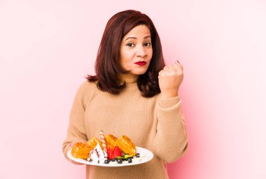 Orta yaşlı Latin bir kadın waffle tutarken izole edilmiş, yumruğunu kameraya gösteriyor, agresif bir yüz ifadesi var..