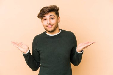 Genç Arap bir adam bej bir arka planda izole edilmiş sorgulama sırasında omuz silkiyor ve şüphe duyuyor..