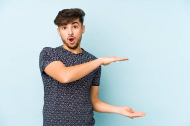 Mavi arka planda izole edilmiş genç bir Arap adam elleri arasında bir kopya alanı tutarken şok olmuş ve hayrete düşmüş..